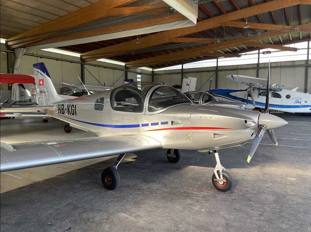 A silver Sonaca 200 aircraft with blue and red stripes, registration HB-KGI, parked inside a hangar.