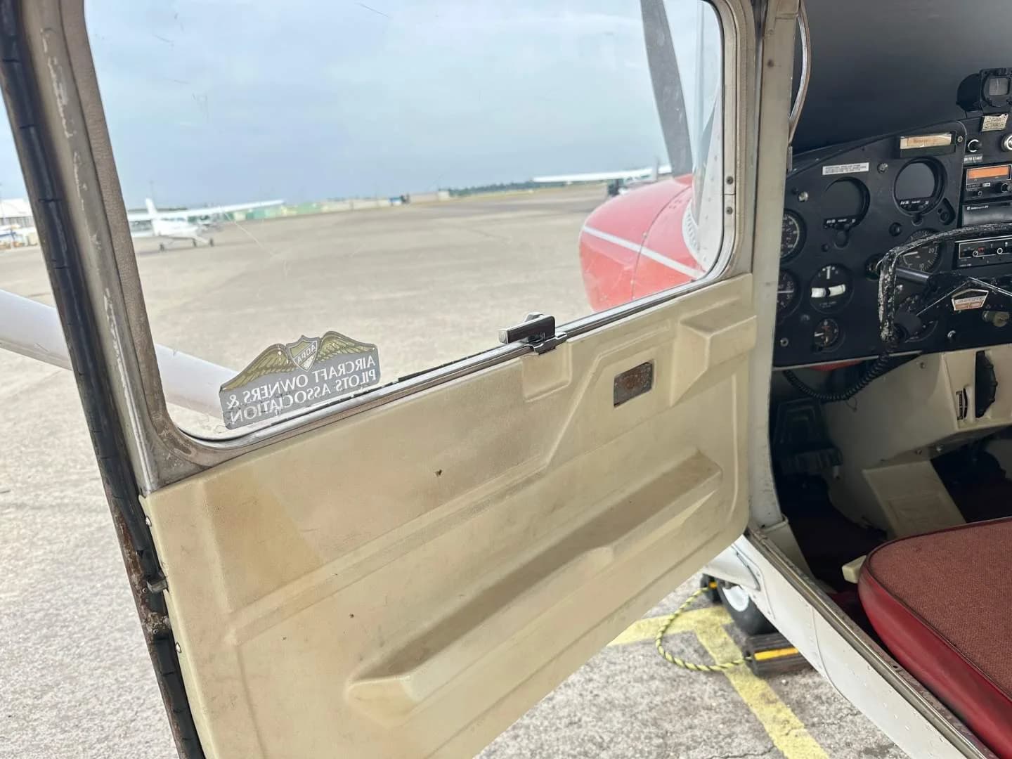 View from outside the open door of the Cessna 150, showing part of the instrument panel and an airfield outside.