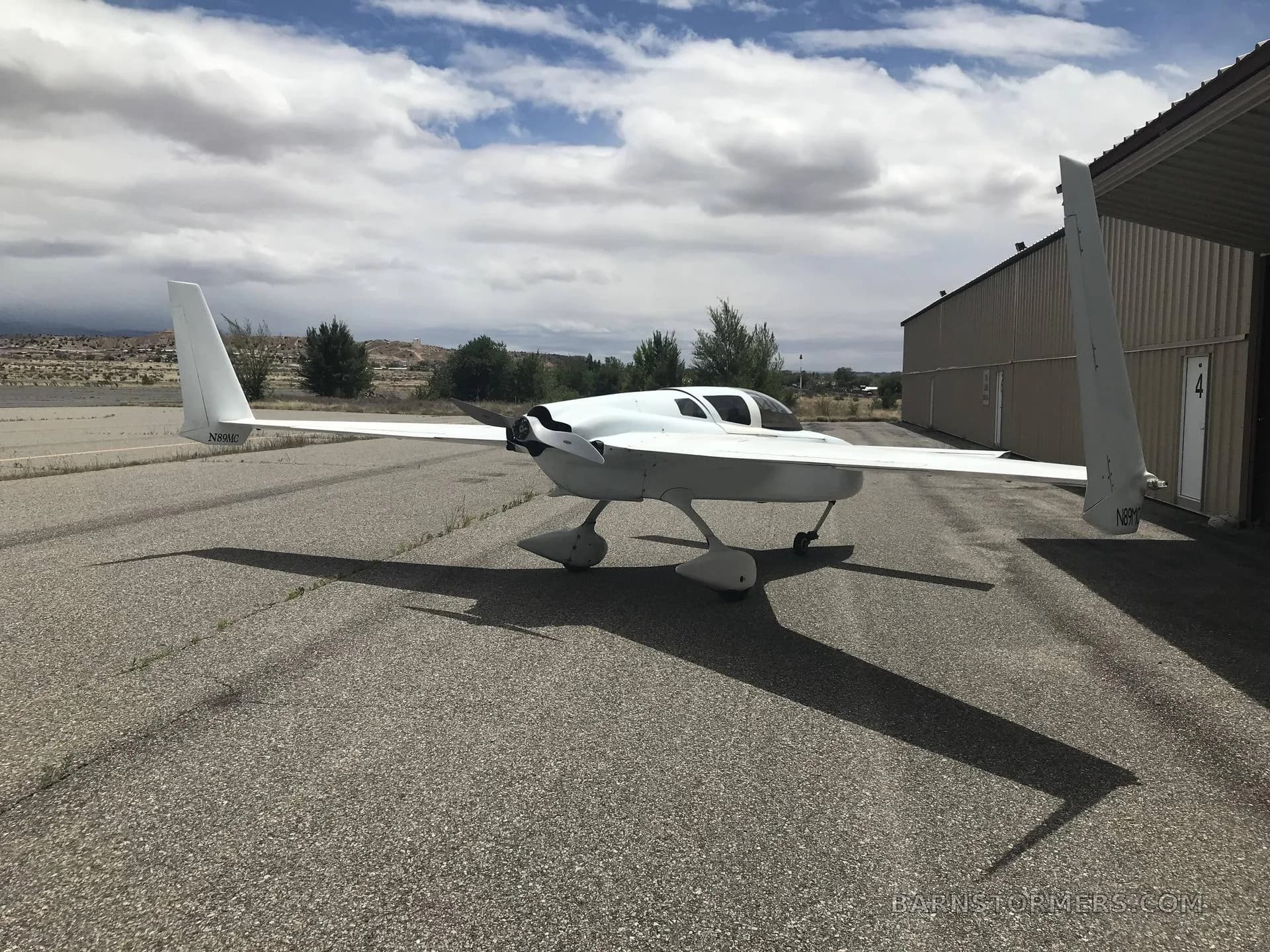 A white, canard-wing aircraft with a pusher propeller is parked on an asphalt runway next to a hangar under a partly cloudy sky.