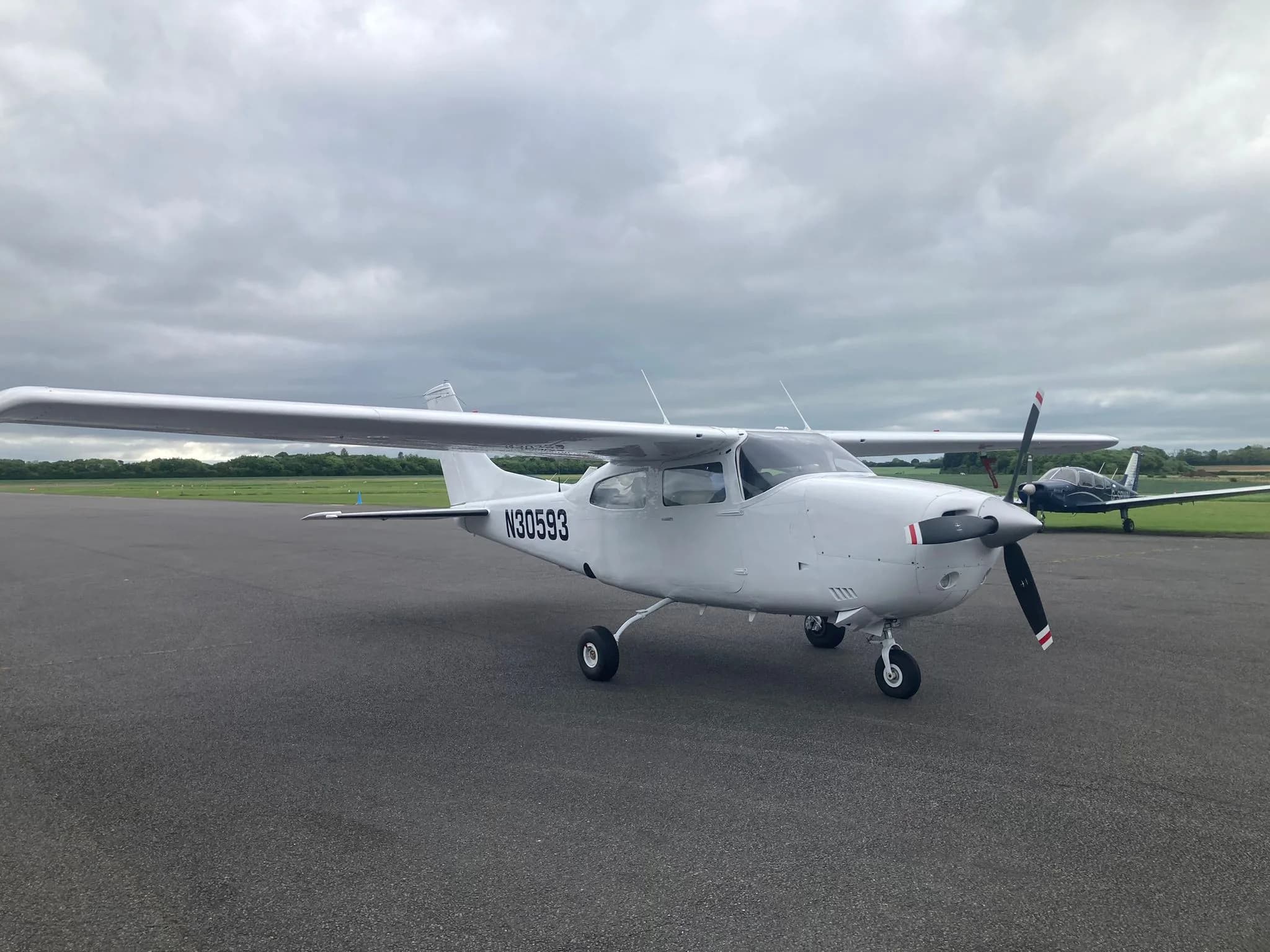 A white Cessna 172 aircraft N30593 seen from the front-left on an airfield with a dark blue aircraft in the distance.