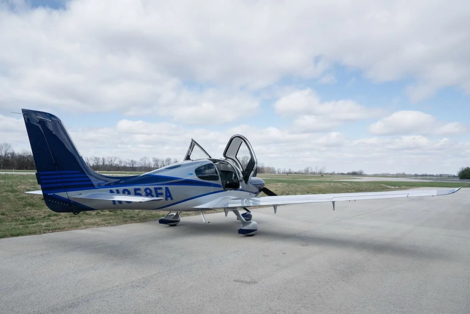 A silver and blue Cirrus SR22T aircraft, registration N358FA, with its gull-wing doors open, parked on an airfield.