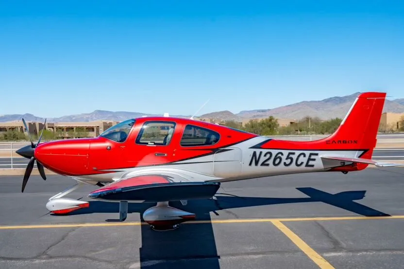 A red and silver Cirrus SR22T GTS Carbon aircraft, registration N265CE, parked on an airfield with mountains in the background.
