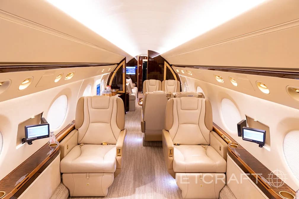 The luxurious interior cabin of a private jet features beige leather seats, individual screens, and warm lighting.