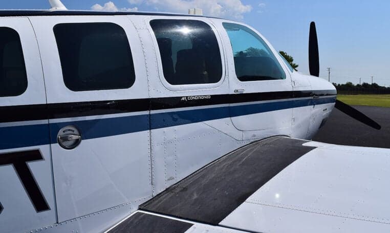 A close-up view of the white and blue fuselage and wing of the Beechcraft Baron aircraft, showing the 'AIR CONDITIONED' label.