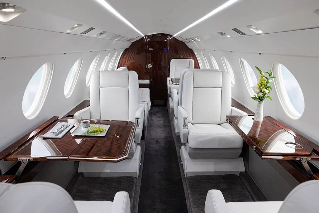 The elegant interior cabin of a business jet with light gray seats, deployed wooden tables, and a floral arrangement.
