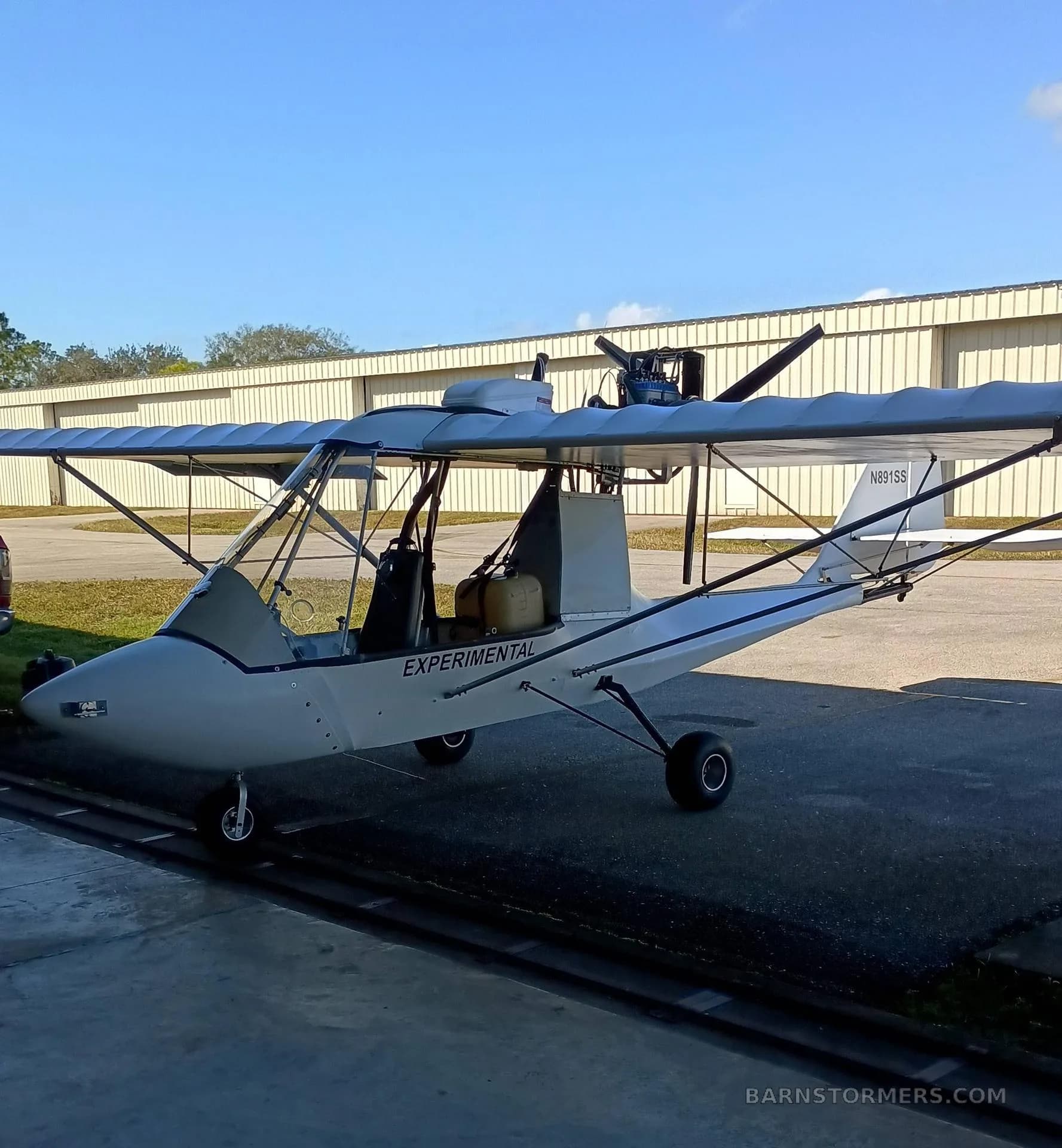 A white experimental ultralight aircraft with a high wing and exposed engine is parked on asphalt in front of a metal hangar, seen from the front-left side.
