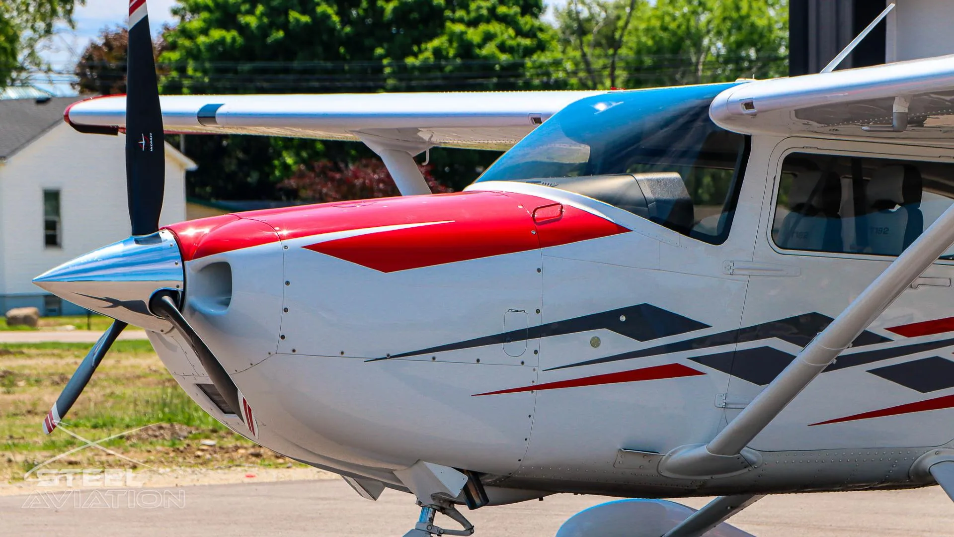 A close-up side view of the nose and propeller of a white Cessna T182T Skylane TC with red and grey stripes, parked outdoors.