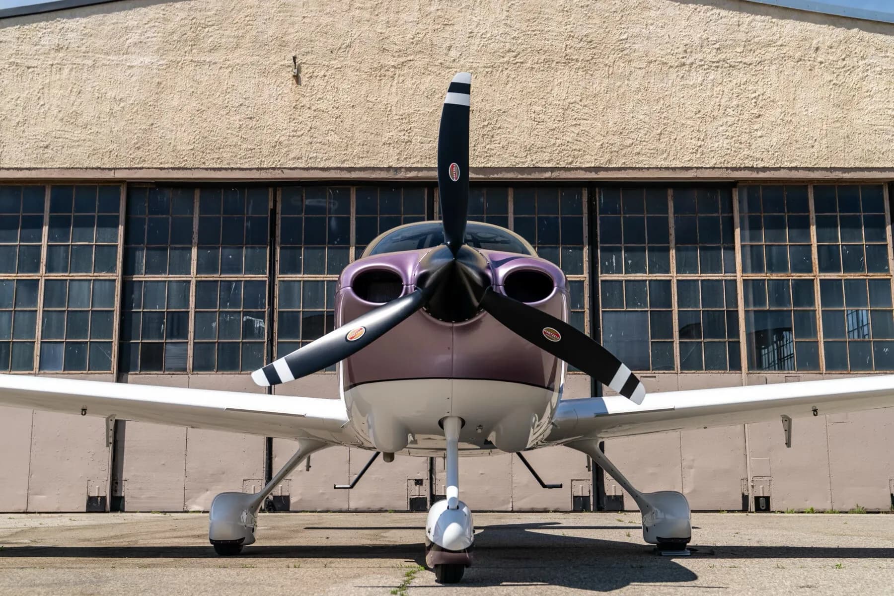 A head-on view of the metallic lavender and white Cirrus SR22 aircraft with its propeller, parked in front of a hangar.