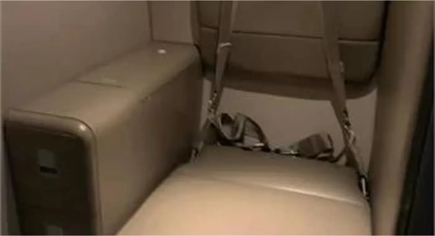 A close-up view of a single beige aircraft seat with a seatbelt next to a matching storage unit.