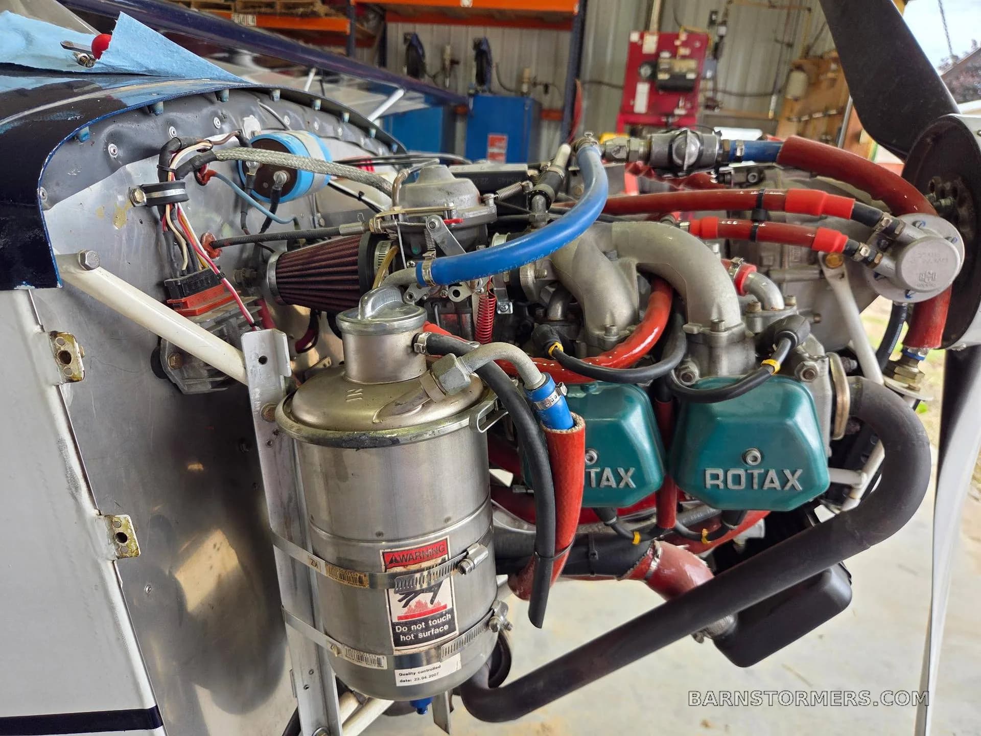 A close-up view of a Rotax aircraft engine with exposed components and red and blue hoses in a hangar.