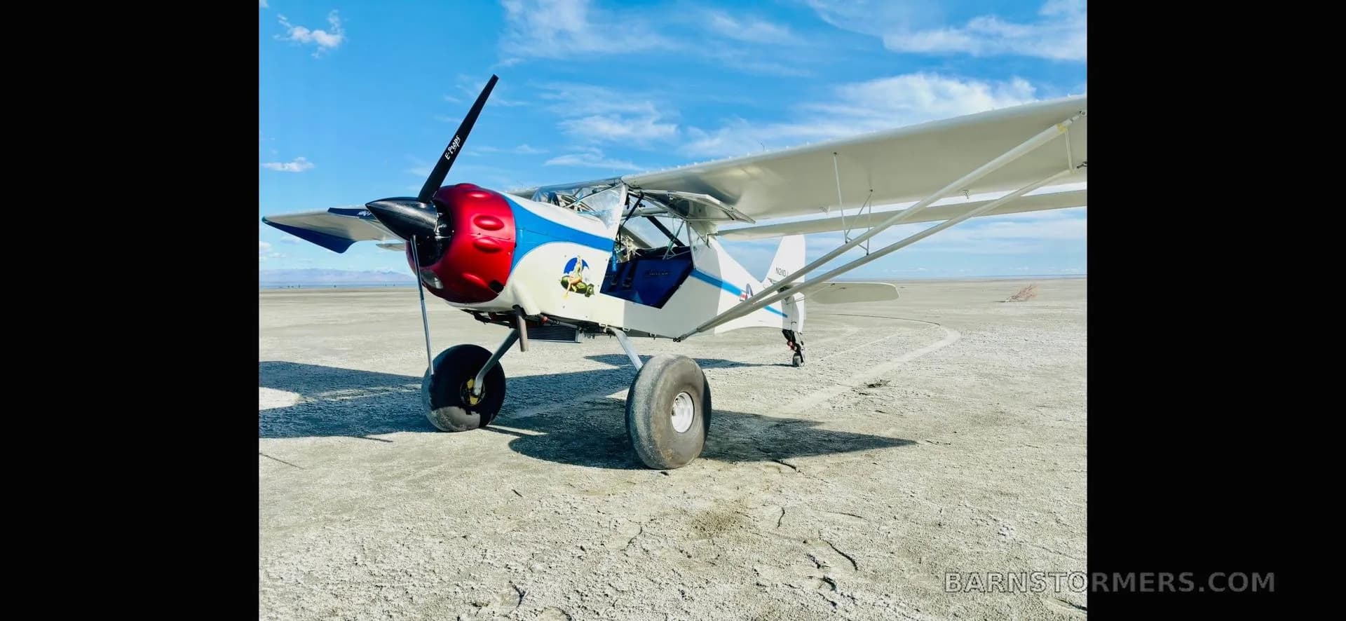 A white Kitfox aircraft with a red and blue cowl and large tires, parked on a vast dry lakebed under a blue sky.