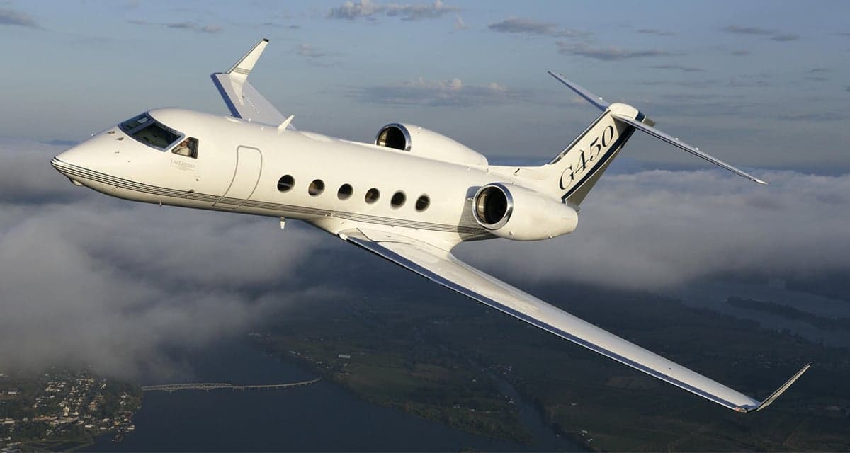 A white Gulfstream G450 business jet with black accents on the tail flies over a landscape with water and a bridge.
