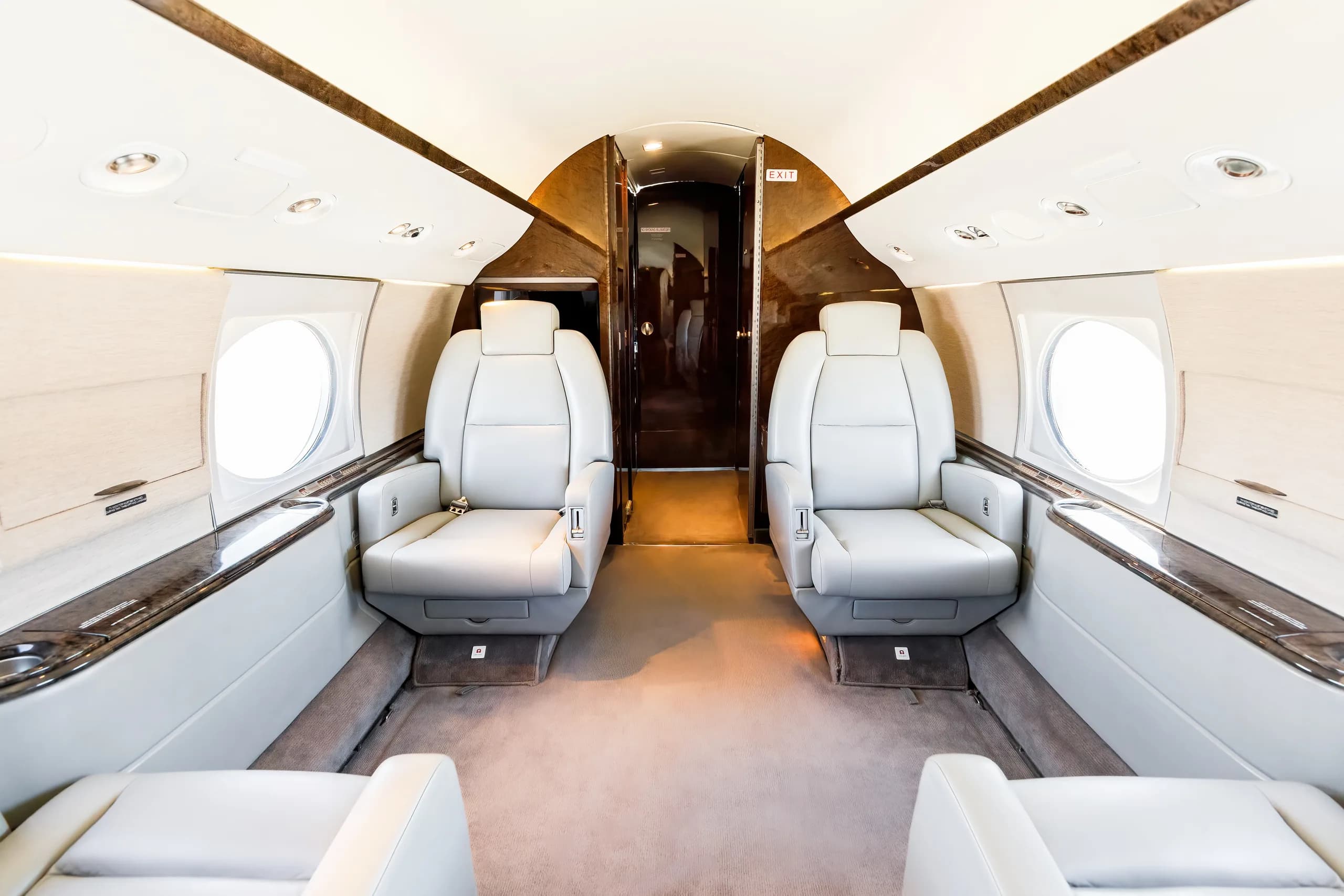 The interior cabin of a private jet features two rows of light gray leather seats facing each other, with a dark wood-paneled rear section.
