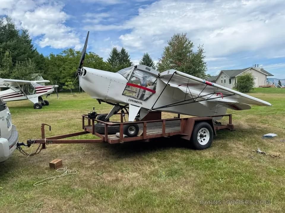 A white experimental aircraft with folded wings loaded onto a utility trailer, hitched to a white SUV, with another aircraft in the background.