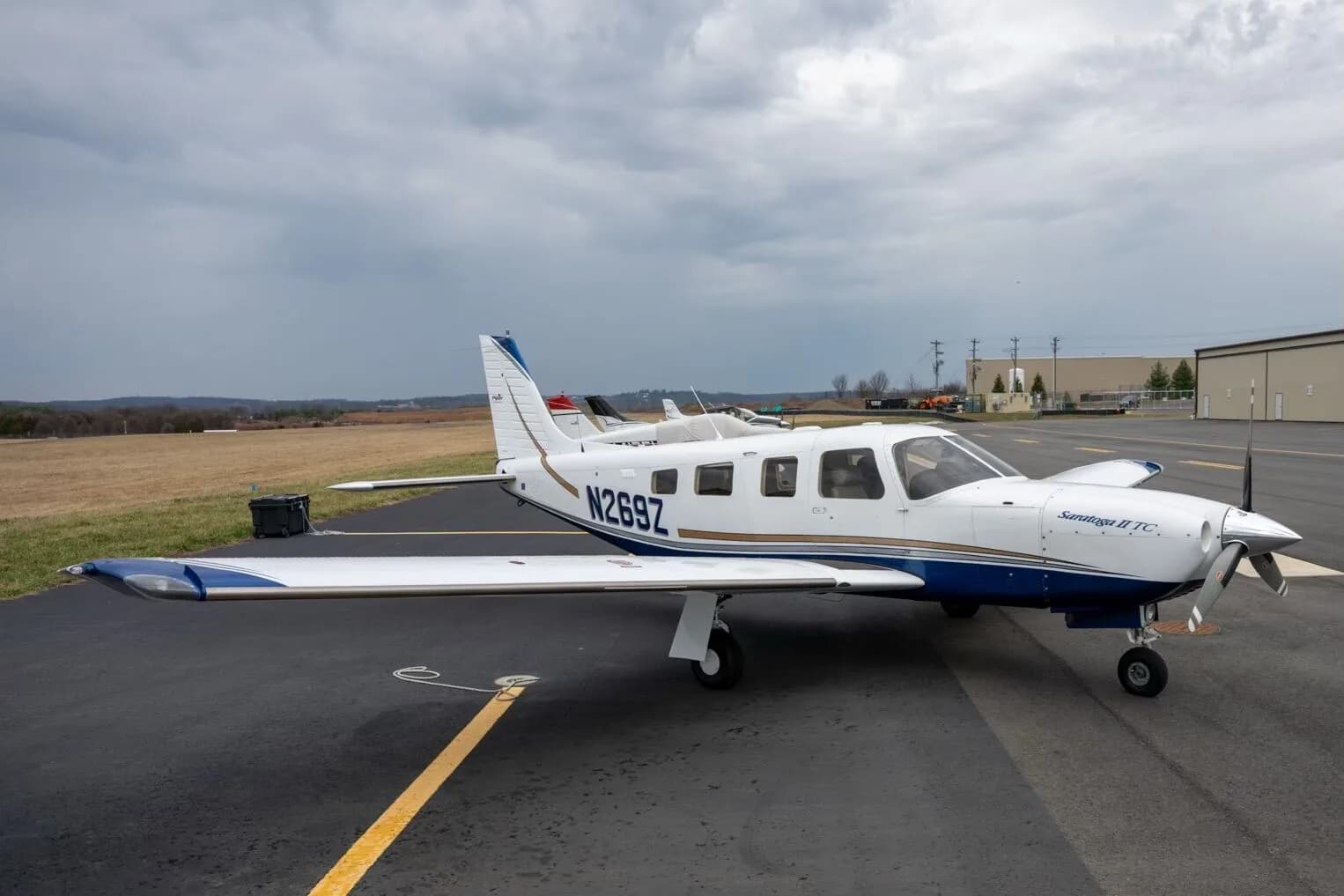 A side view of a white Piper Saratoga II TC aircraft, tail number N269Z, parked on the tarmac with a field and buildings in the distance.