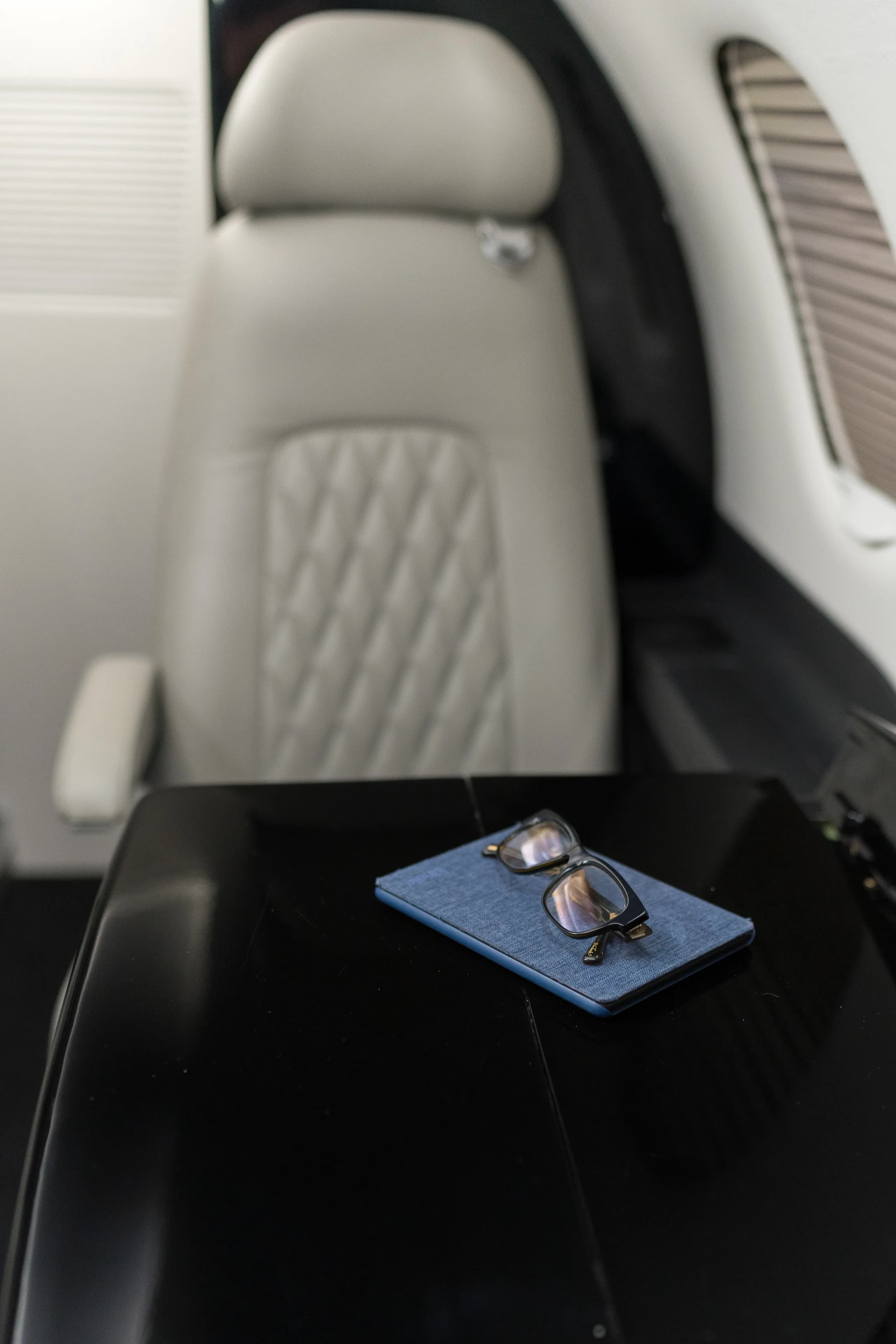 A close-up of a light gray leather cabin seat with a quilted pattern and a black table with eyeglasses and a blue case.