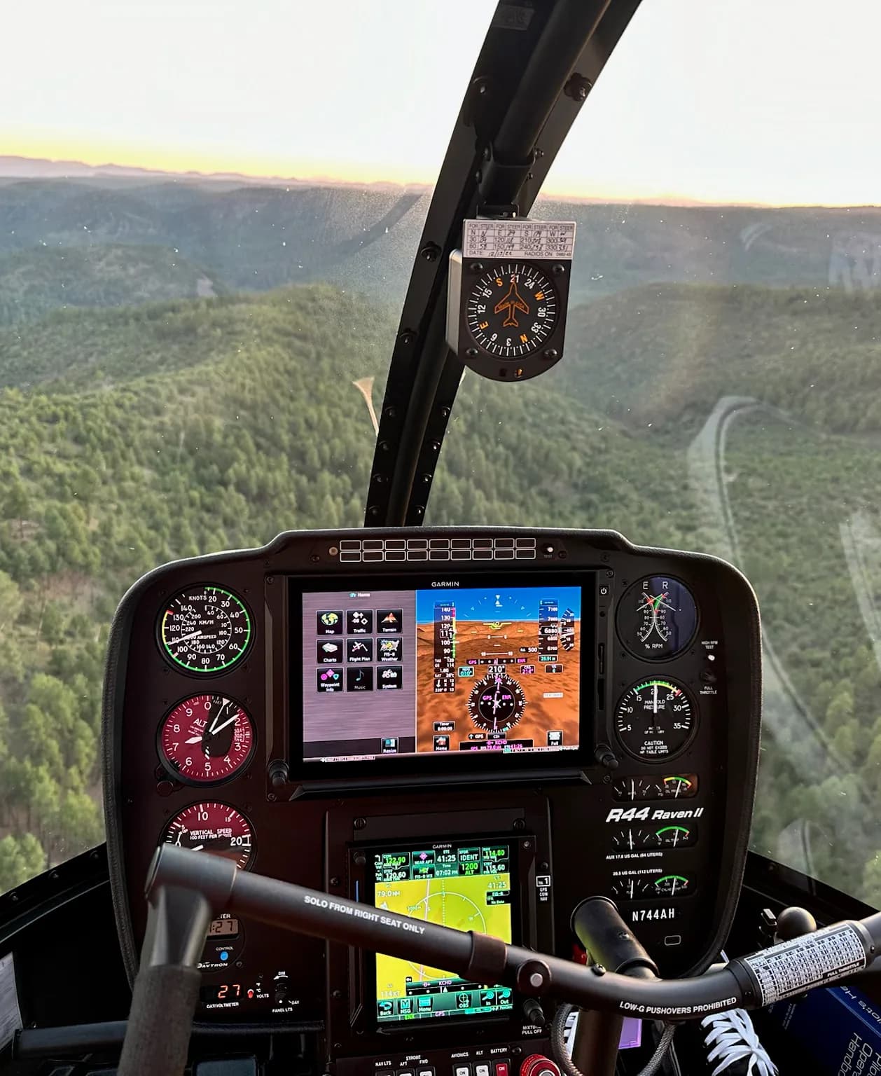 The cockpit of a Robinson R44 Raven II helicopter with a view of a forested mountain landscape at sunset.