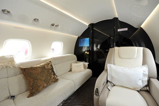 A section of a private jet's interior cabin featuring a cream-colored sofa, a single seat, and a small screen.