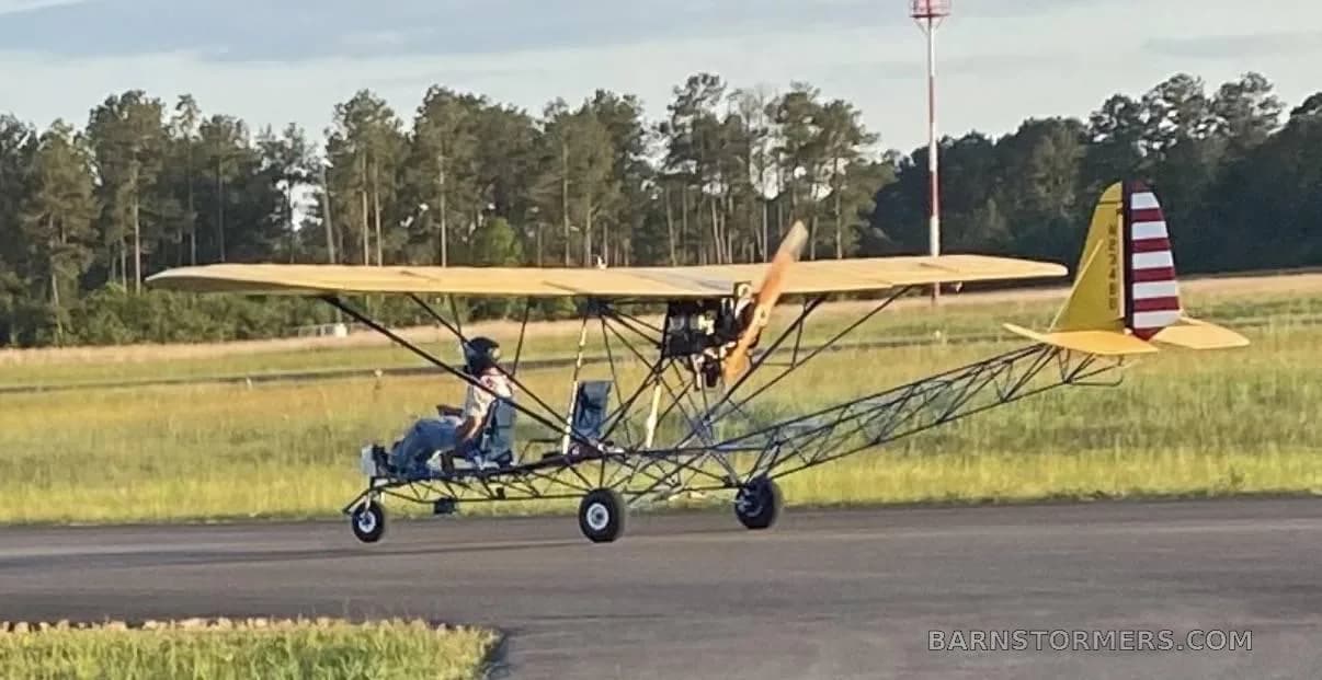A yellow-winged, blue-framed ultralight aircraft with a pilot is taxiing on an asphalt runway with trees in the background.