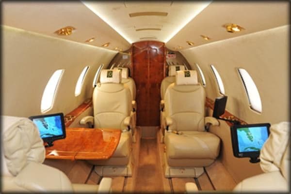The main cabin of a private jet features multiple cream leather seats, individual entertainment screens, and polished wood tables.