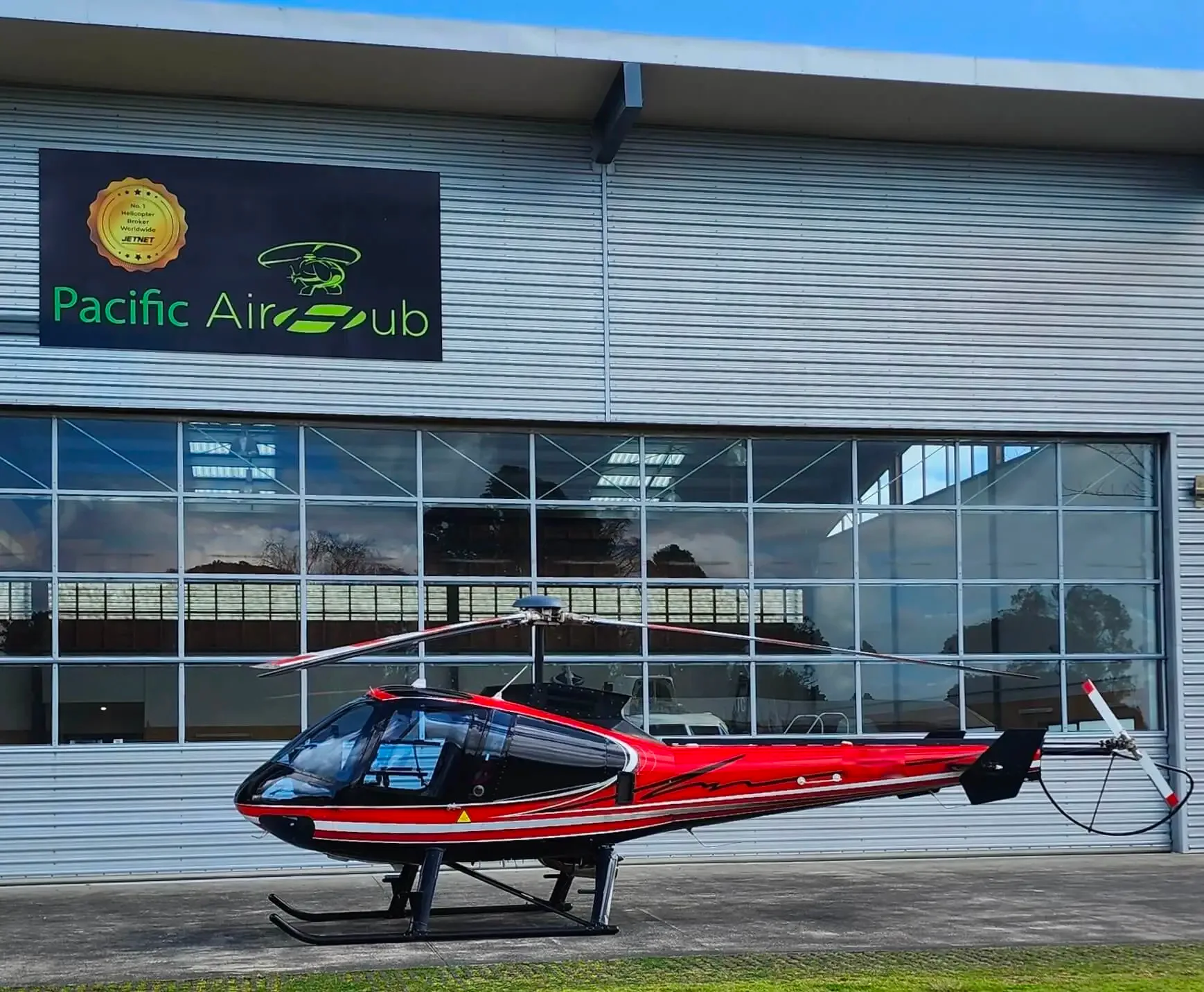 A red and black helicopter is parked on a tarmac in front of a modern hangar with a "Pacific Air Hub" sign.