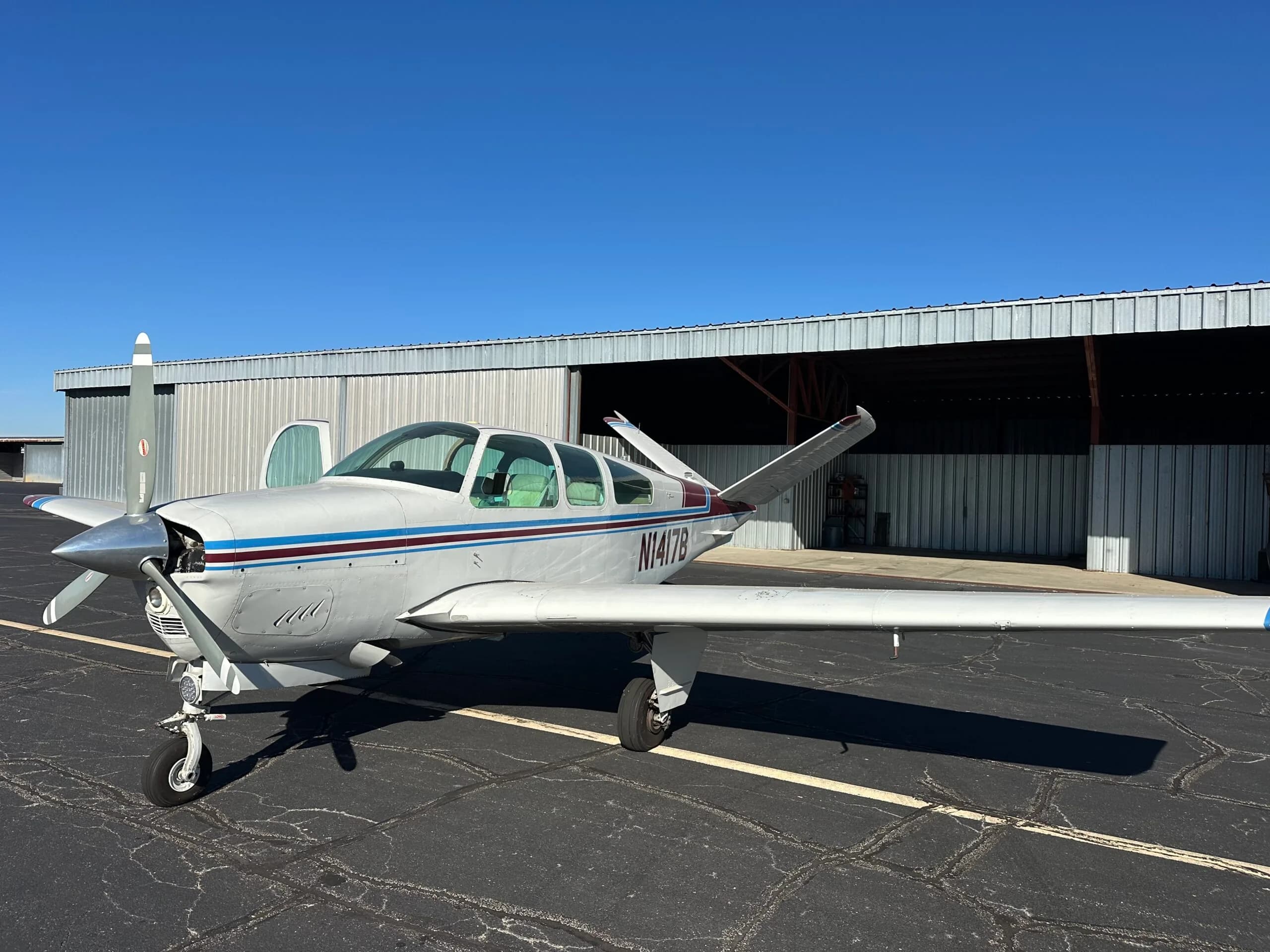 A white Beechcraft Bonanza with blue and maroon stripes is parked on a tarmac in front of an open hangar on a sunny day.