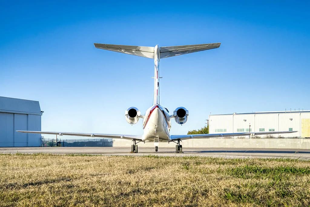A white business jet (N24KT) is viewed from the rear, parked on a tarmac with hangars in the background.