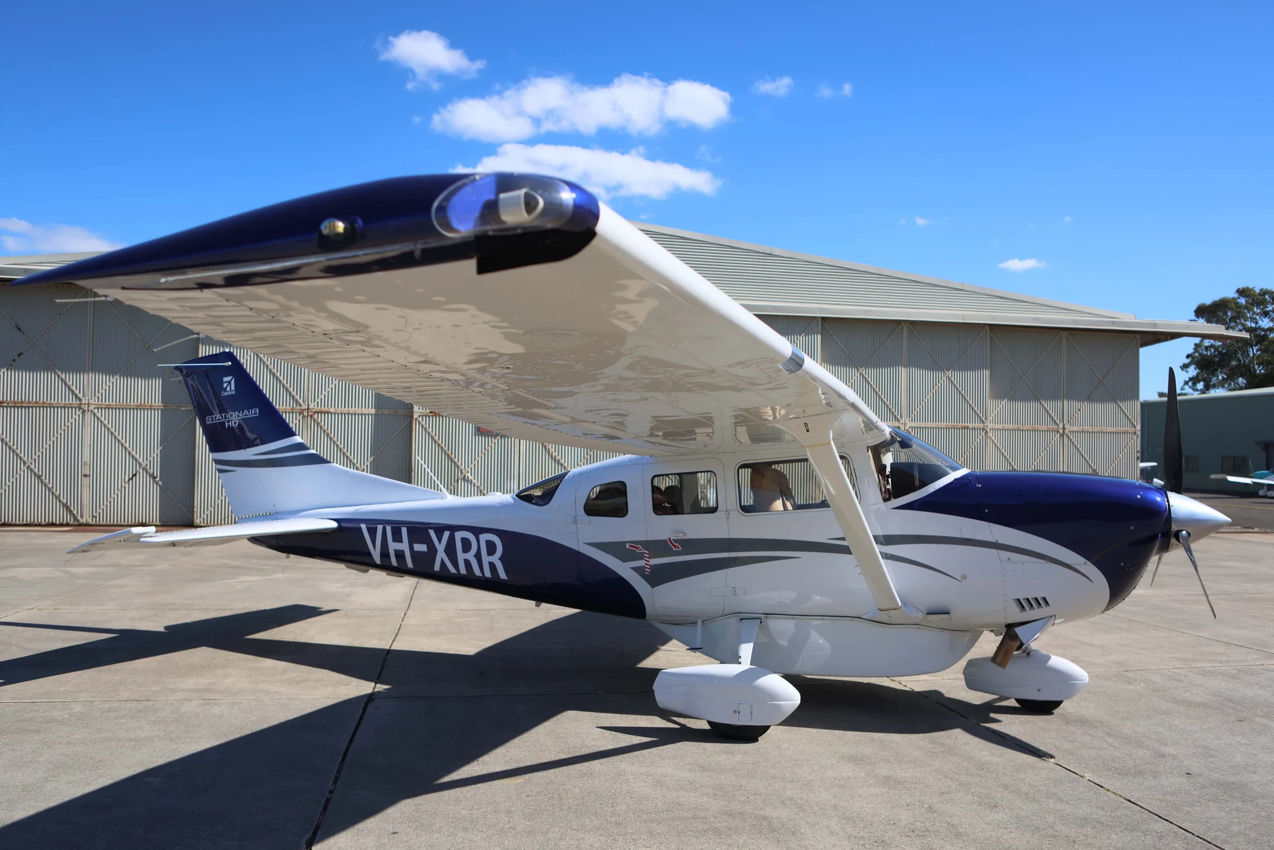 A white and blue Cessna 206 Stationair HD aircraft with registration VH-XRR is parked on an airfield in front of a hangar under a blue sky.