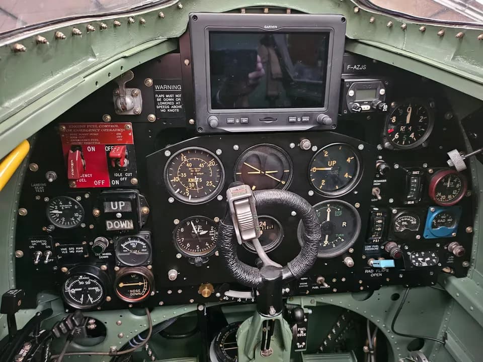 The instrument panel of a Supermarine Spitfire cockpit, showing various gauges, switches, and a modern Garmin GPS unit, with F-AZJS visible.