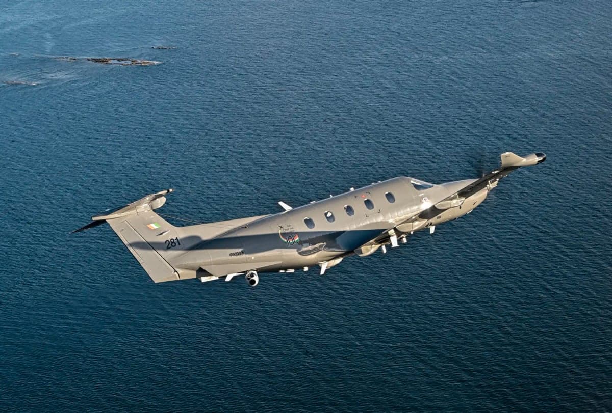 A Pilatus PC-12 aircraft in military livery flies over the ocean.