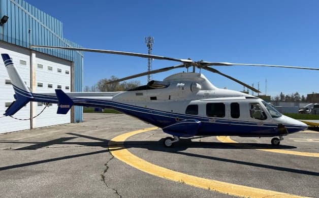 A white and blue helicopter sits on the tarmac in front of a hangar.