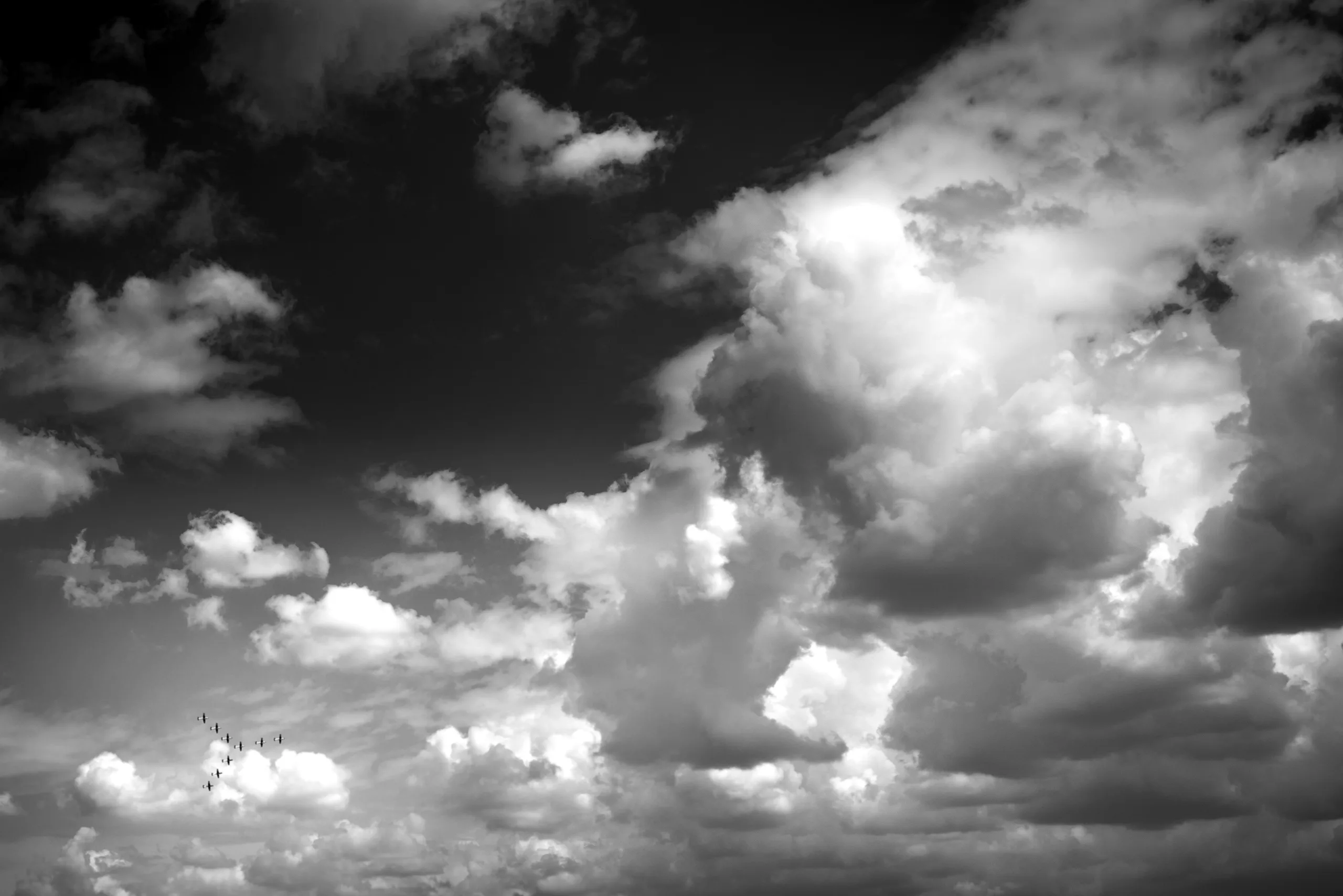 A black and white photo of a cloudy sky with a small formation of aircraft flying in the distance.
