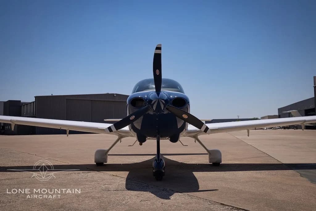 A head-on view of the white Cirrus SR22 aircraft with a dark blue nose and propeller, parked on an airport tarmac.