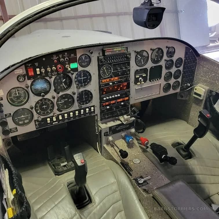 A wide view of an aircraft cockpit, showing the instrument panel, control sticks, and light grey quilted seats.