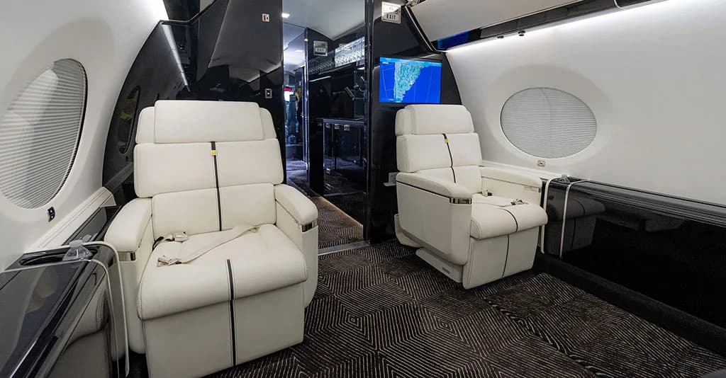 Two white leather seats in the business jet cabin, with a TV screen visible in the background.