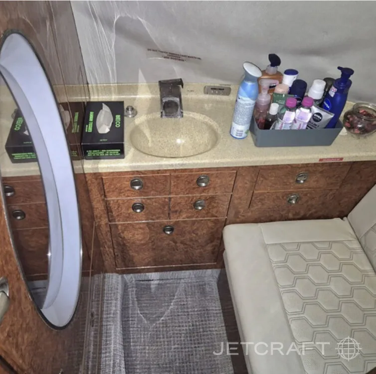 The lavatory or galley area of a private jet, featuring a sink, mirror, wood cabinetry, and various toiletries.