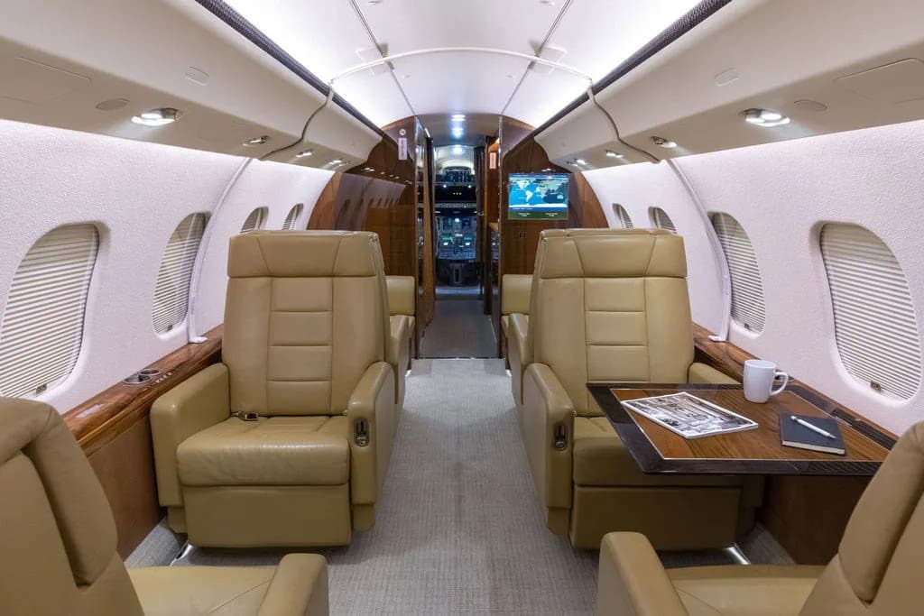 A view down the business jet cabin showing tan leather seats, a table, and a monitor at the front.