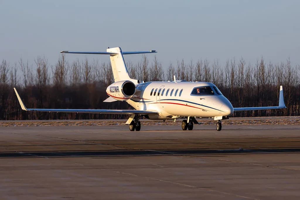A white Learjet 60, registration N221WP, with red and blue stripes, is seen on an airport tarmac with trees in the background.