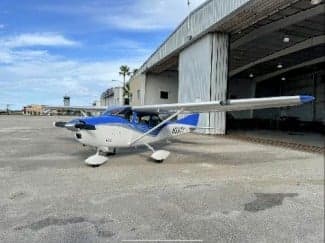 A Cessna 182 Skylane aircraft with a blue and white paint scheme is parked on the tarmac in front of a hangar.