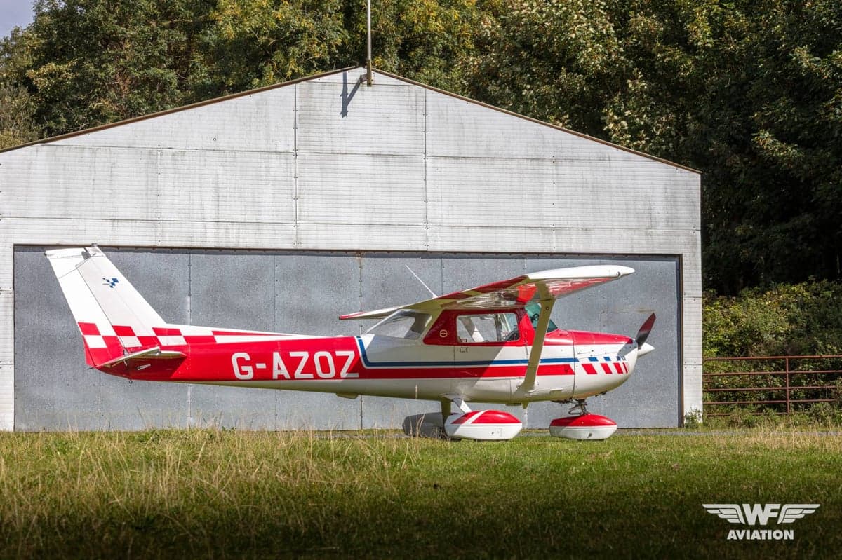 A red and white Cessna 150 Aerobat (G-AZOZ) with a checkered tail, parked on grass in front of a hangar.