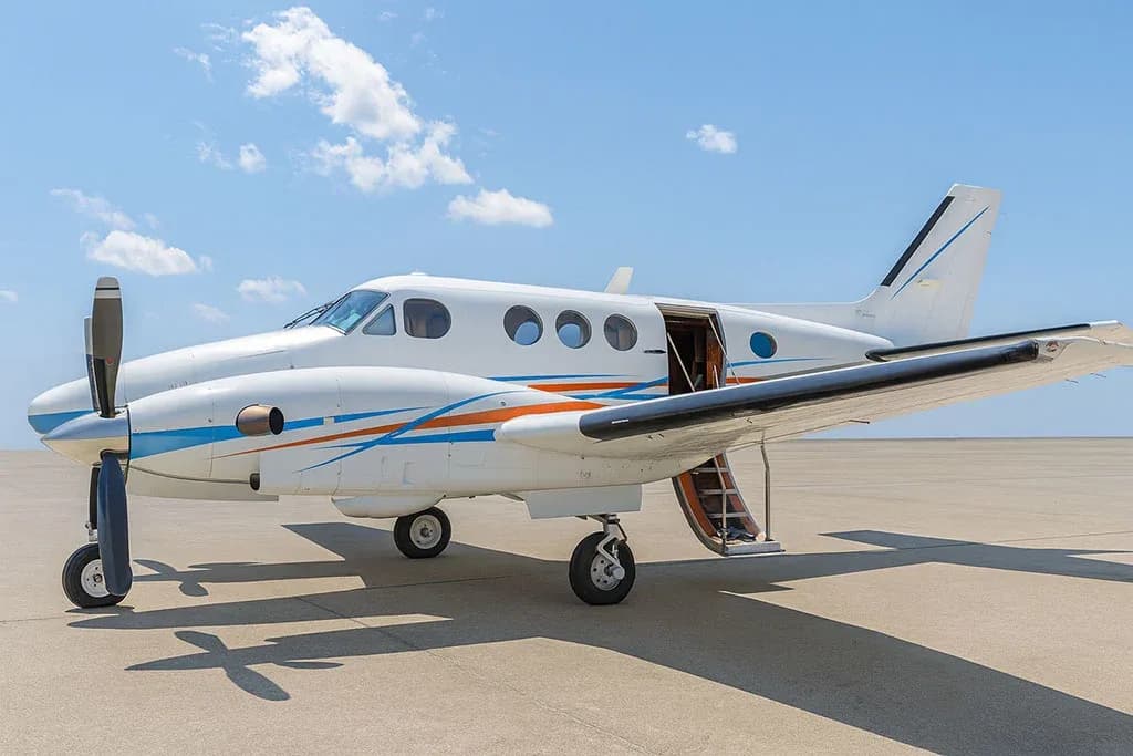 A white twin-engine turboprop aircraft with blue and orange stripes on the fuselage and wings, parked on a tarmac under a blue sky.