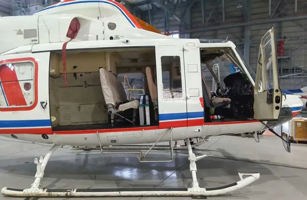 The side of a white helicopter with red and blue stripes, showing open cabin and cockpit doors in a hangar.
