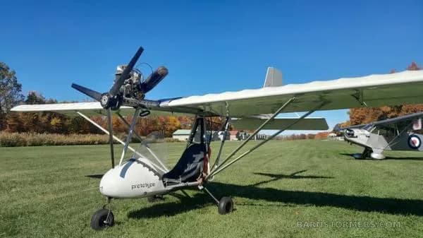 A white ultralight aircraft with 'prototype' on its nose is parked on a grassy field, with another aircraft visible in the background.