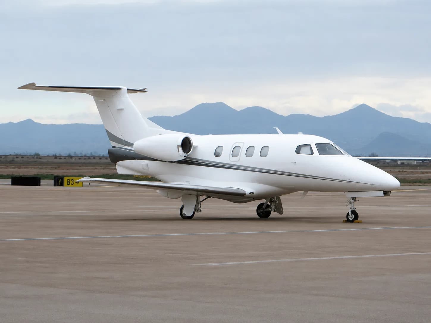 A white business jet with grey stripes is parked on an airport tarmac with mountains in the background under a cloudy sky.