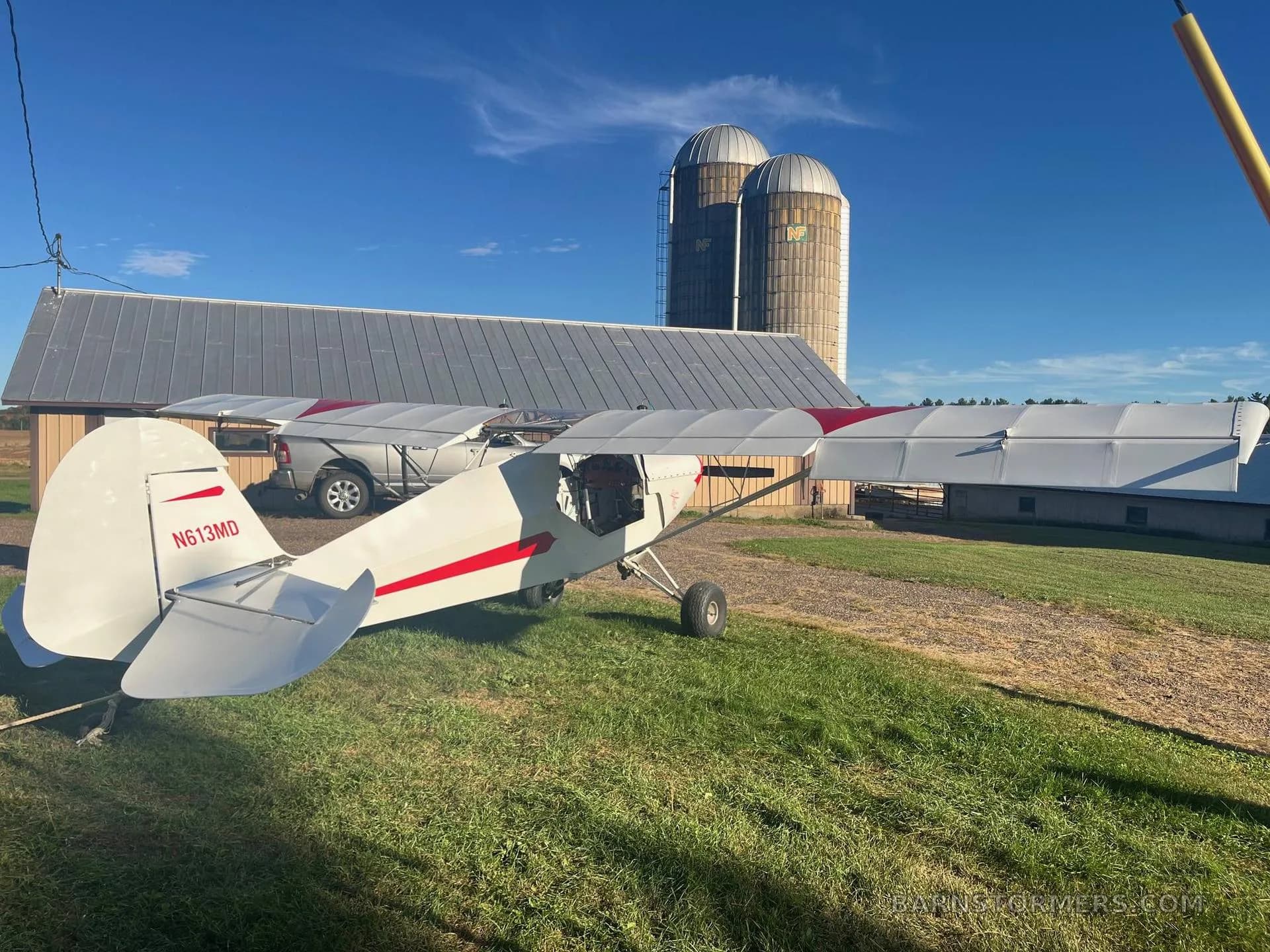 Rear-side view of a white and red experimental aircraft with tail number N613MD, parked on grass with silos and a barn in the background.