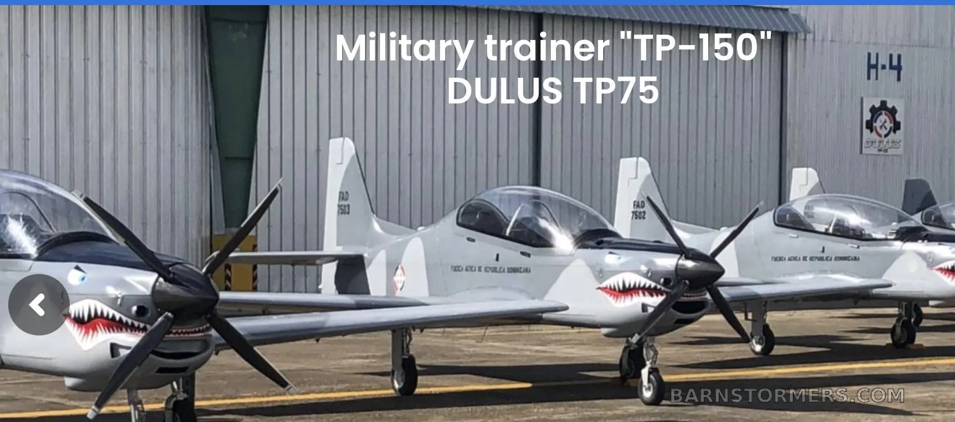 A line of DULUS TP75 military trainer aircraft in light gray camouflage with shark mouth nose art, parked in front of a hangar.