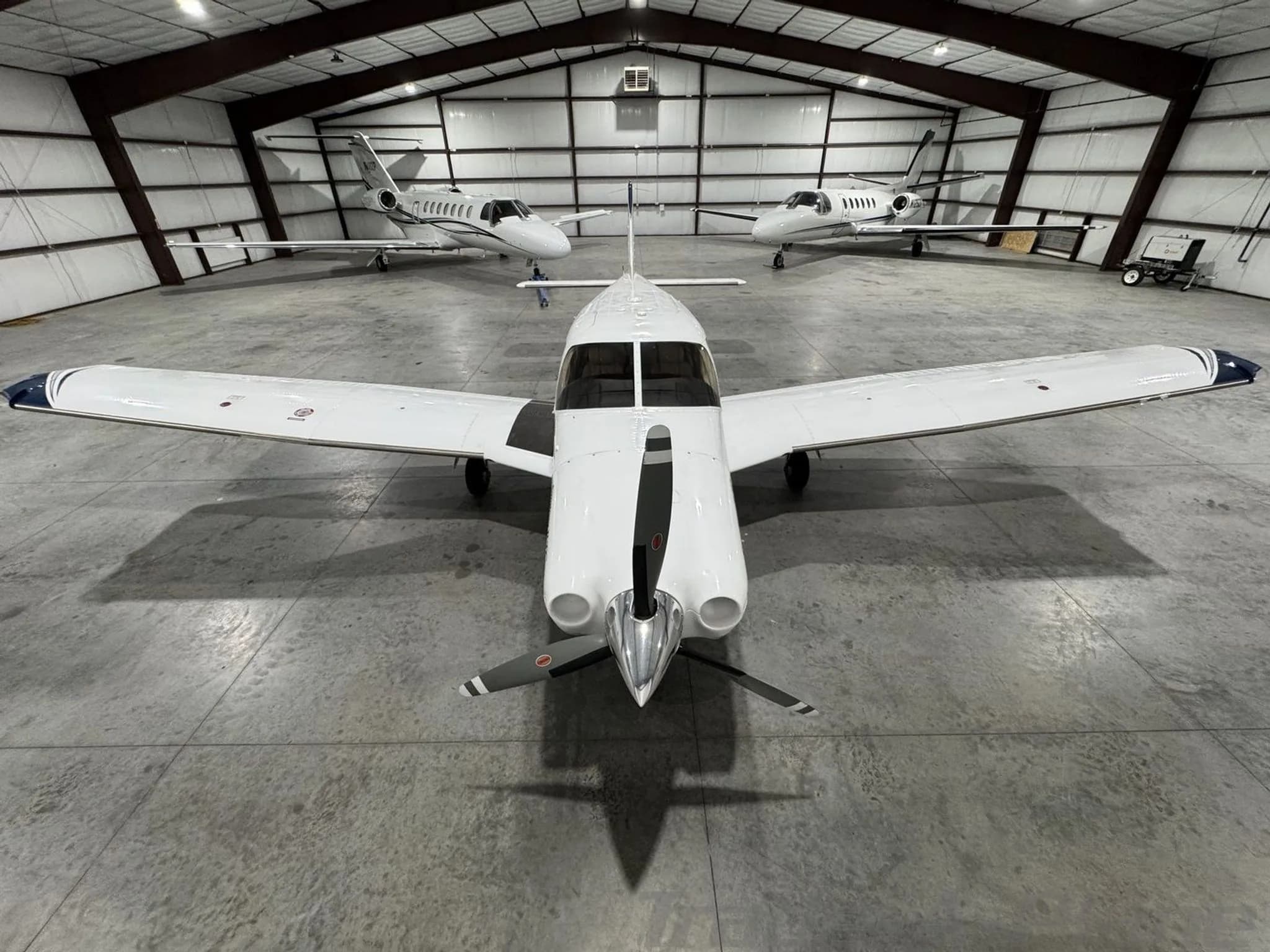 A head-on view of the Piper Saratoga II TC N436MA with its propeller, parked in a hangar between two jets.