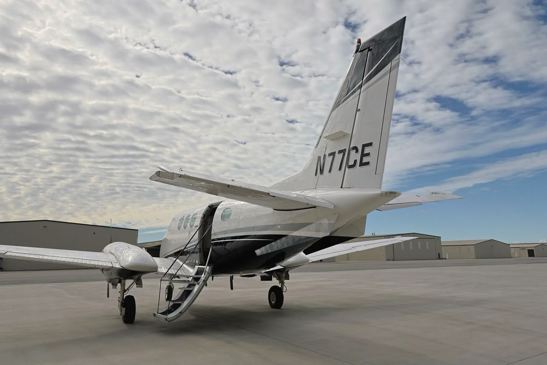 A rear-left quarter view of the N77CE twin-engine turboprop aircraft with its passenger door open and stairs deployed.