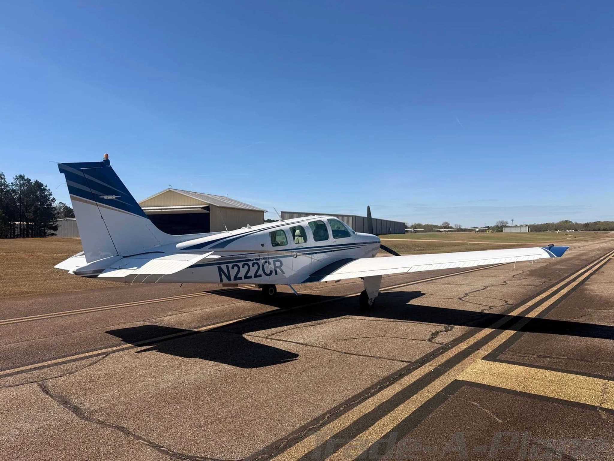 A white Beechcraft Bonanza N222CR with blue and gold stripes parked on an asphalt runway with a hangar in the background.