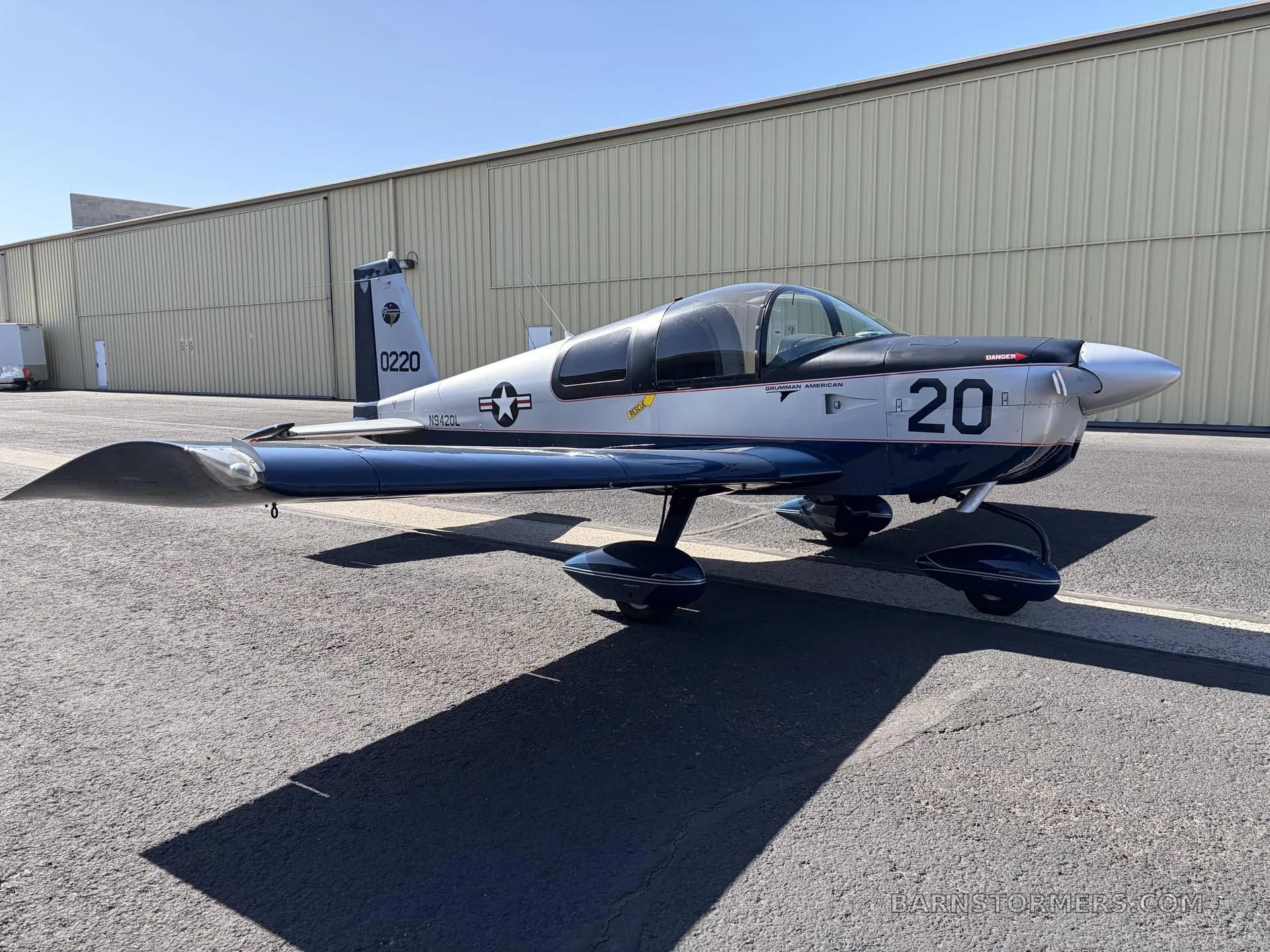 A silver and blue Grumman American AA-5 Traveler aircraft with a military-style paint scheme, parked on an asphalt ramp next to a hangar.
